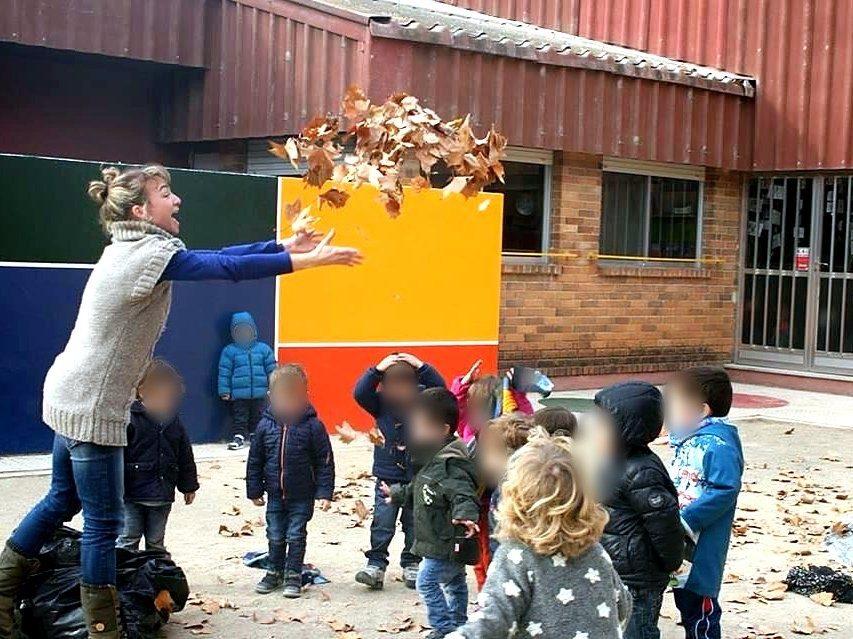 Maestra jugando con las hojas de otoño junto a un grupo de peques en el patio